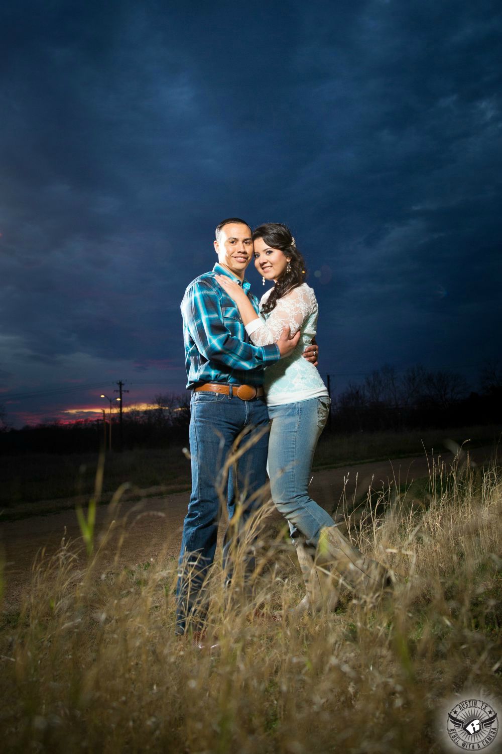 Brunette Latino girl with a curly pony tale and dangling earrings in a white lace blouse, blue jeans and tan cowboy boots embraces a Mexican guy with short black hair wearing an aqua and blue plaid flannel long sleeve shirt and dark blue jeans stand together in a golden grassy field at dusk in front of a big bluish cloudy sky and a hint of orange sunset at Mueller Lake Park in this stunning engagement photo in Austin Texas. 